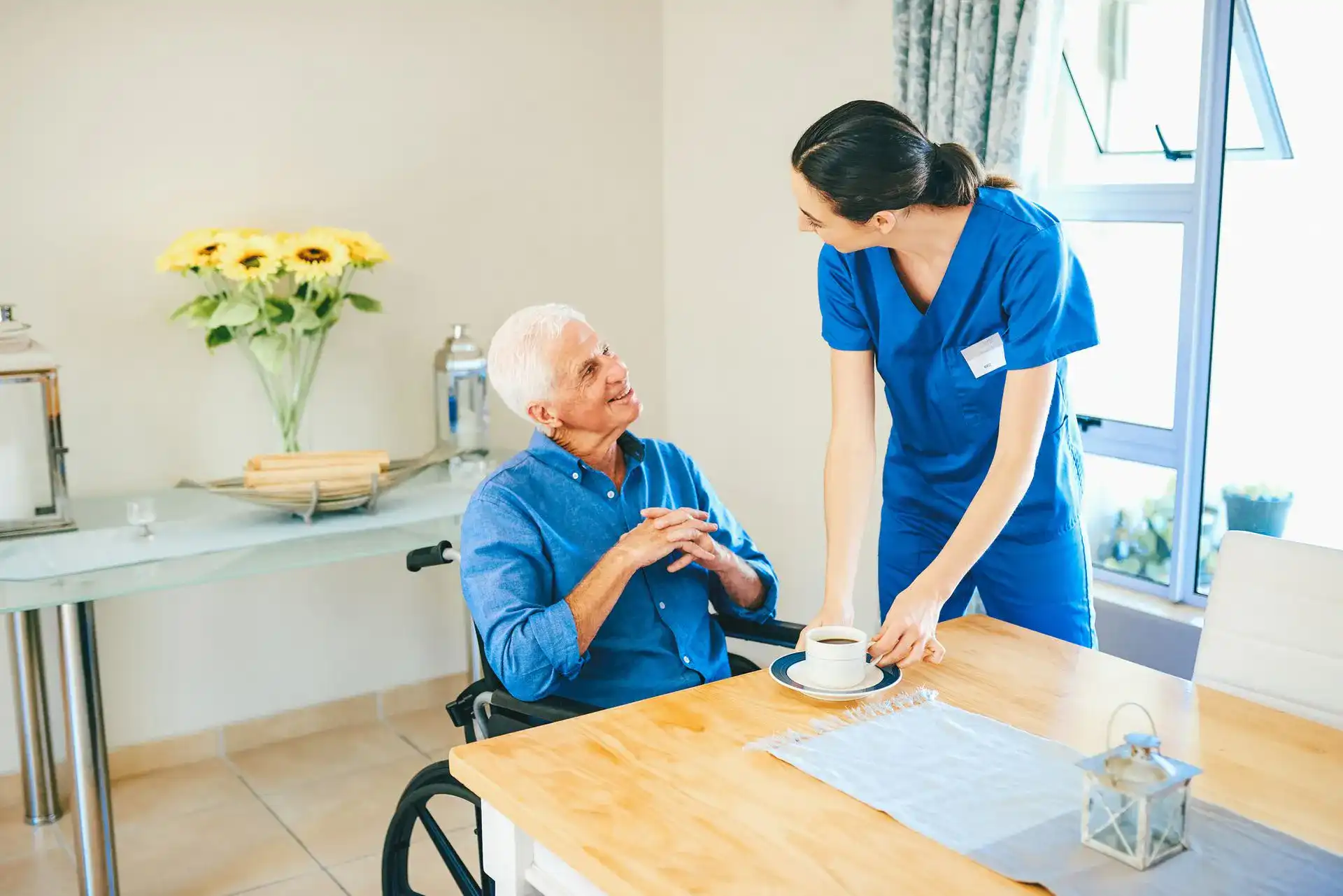 A bright and airy indoor scene showing a caring interaction between an elderly man in a wheelchair and a young female nurse. The man, wearing a blue button-down shirt, is seated at a light-colored wooden table and looking up with a smile at the nurse. The nurse, dressed in professional blue medical scrubs with a name tag, is leaning over to place a cup of coffee on the table in front of him. In the background, there's a vase of bright yellow sunflowers on a console table and a large window that lets in plenty of natural light, creating a warm, positive, and comforting atmosphere.