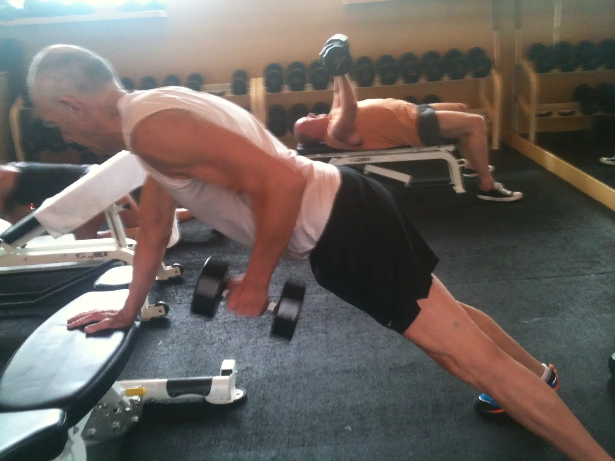 An indoor gym scene featuring several men working out. In the foreground, a man in a white tank top and black shorts is performing a single-arm dumbbell row while leaning on a weight bench. In the background, another man in an orange shirt is lying on a bench performing a chest press with a dumbbell. A rack of various dumbbells lines the wall in the background. The photo is a candid action shot with some motion blur and natural lighting.