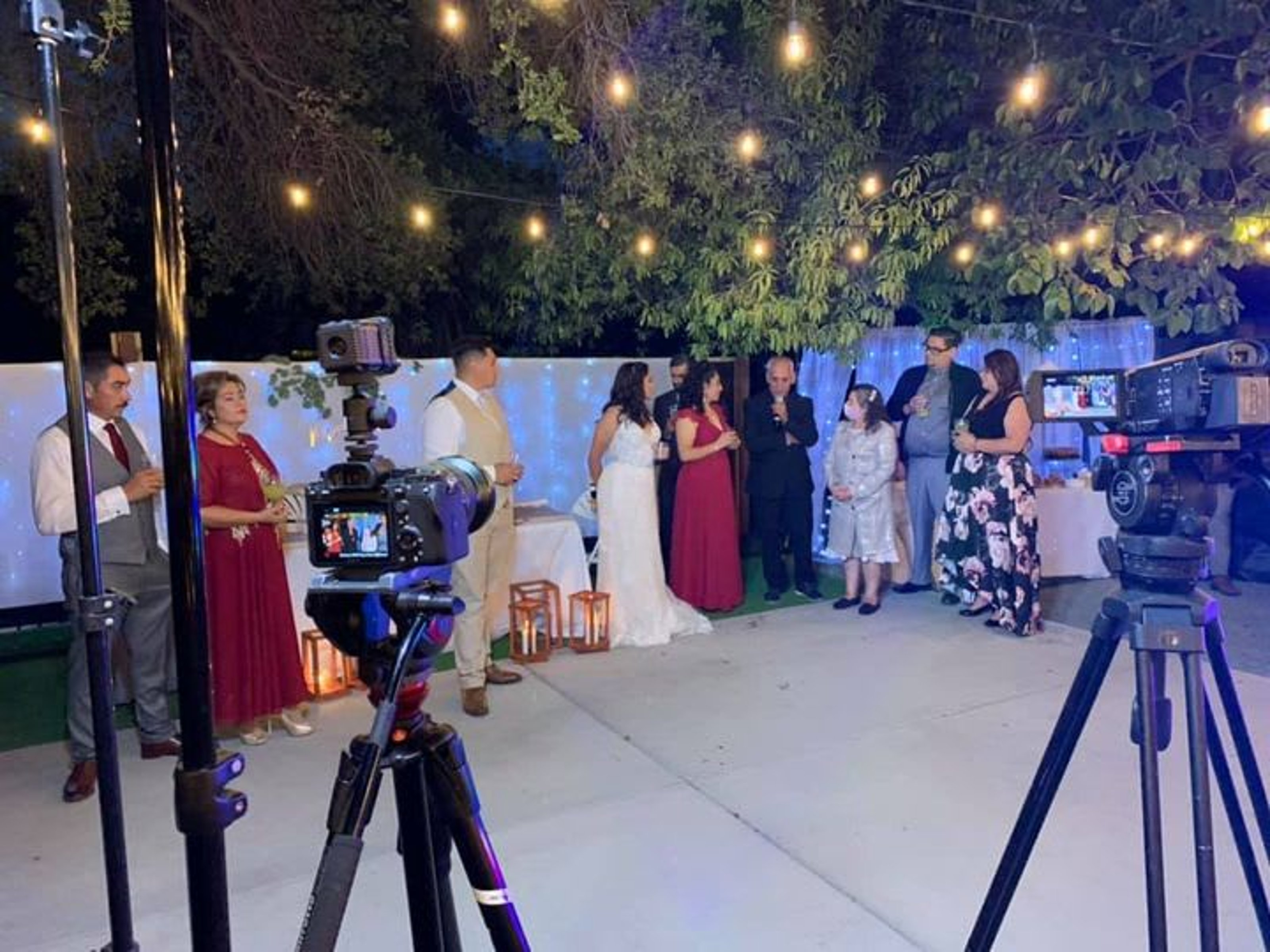 An outdoor evening scene showing a group of people gathered for an event, likely a wedding or formal celebration. In the foreground, two professional video cameras on tripods are positioned to film the group. The setting features warm string lights overhead, a white fence decorated with blue fairy lights, and leafy trees. The participants are dressed in formal attire, including a woman in a white gown and others in suits and dresses. The atmosphere is celebratory and festive, captured from a behind-the-scenes perspective.