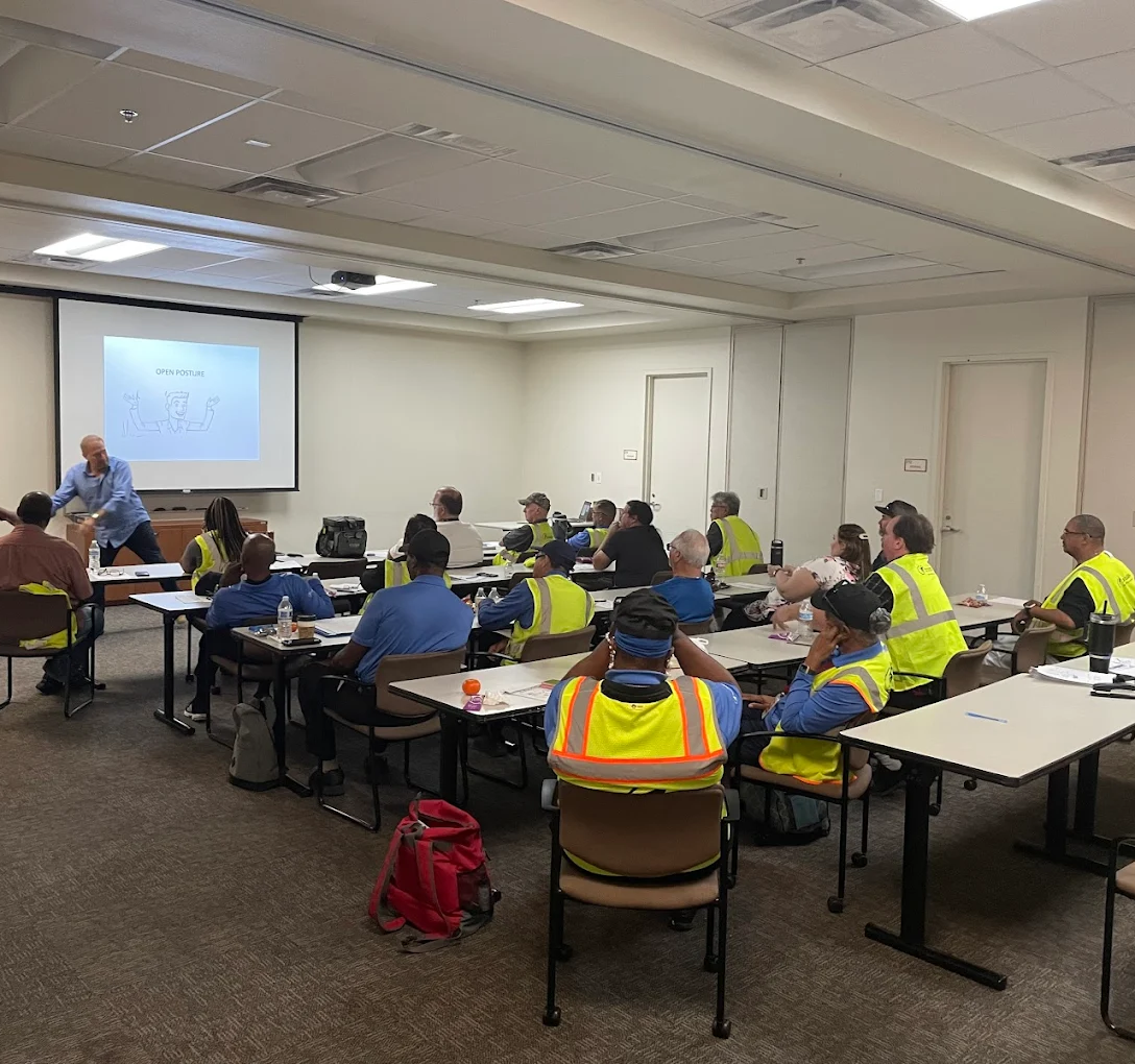 Workers in safety vests attend a training session with an instructor demonstrating "open posture."