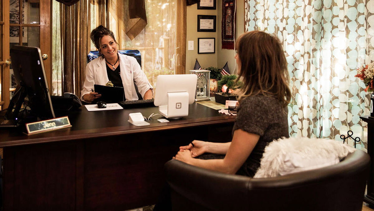 A woman wearing a white medical coat sits behind a dark wooden desk, smiling warmly at a client or patient who is seated in the foreground with her back partially to the camera. The office has a professional yet cozy atmosphere, with green walls, framed certificates, a small bonsai tree, and teal patterned curtains in the background. A computer monitor and a white Square payment terminal are visible on the desk, alongside a nameplate that reads 'Jenn'. The lighting is soft and warm, creating a welcoming and trustworthy mood.